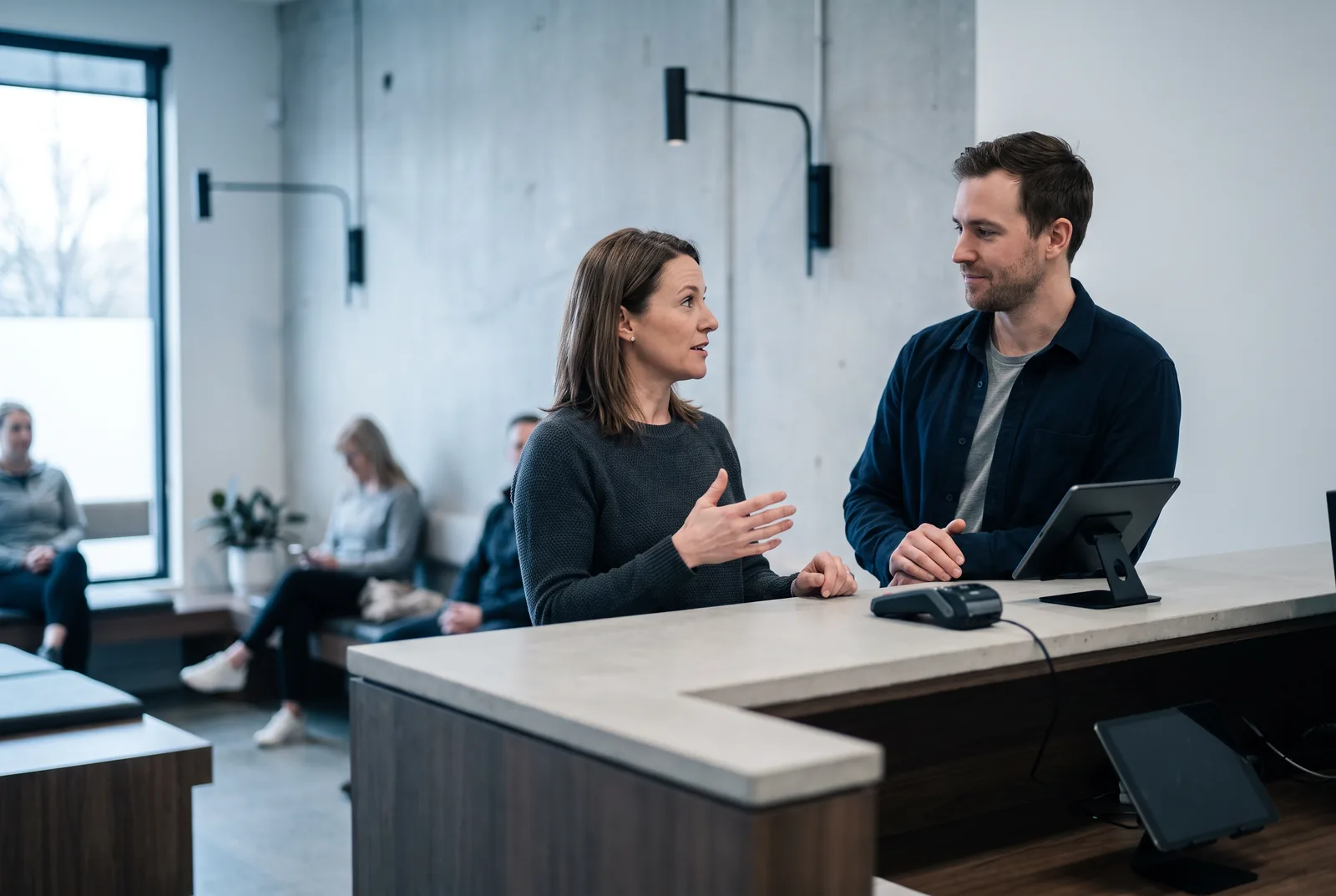 A clinic's operations manager talking with her technical partner at the front desk of a physiotherapy clinic, patients waiting in the background.